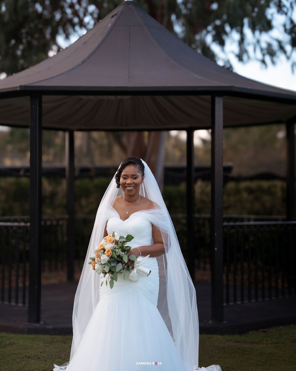 A bride in a white wedding dress poses outdoors under a dark gazebo during a wedding photoshoot.