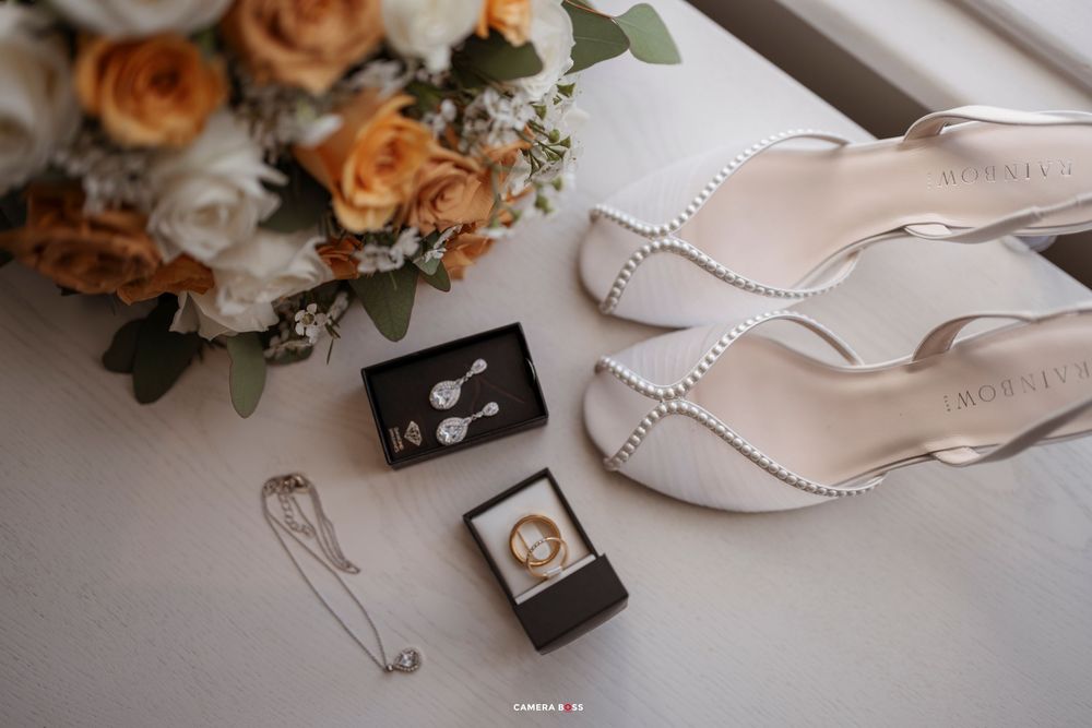 A romantic wedding detail shot featuring peach roses, white shoes, and delicate jewelry on a light background.