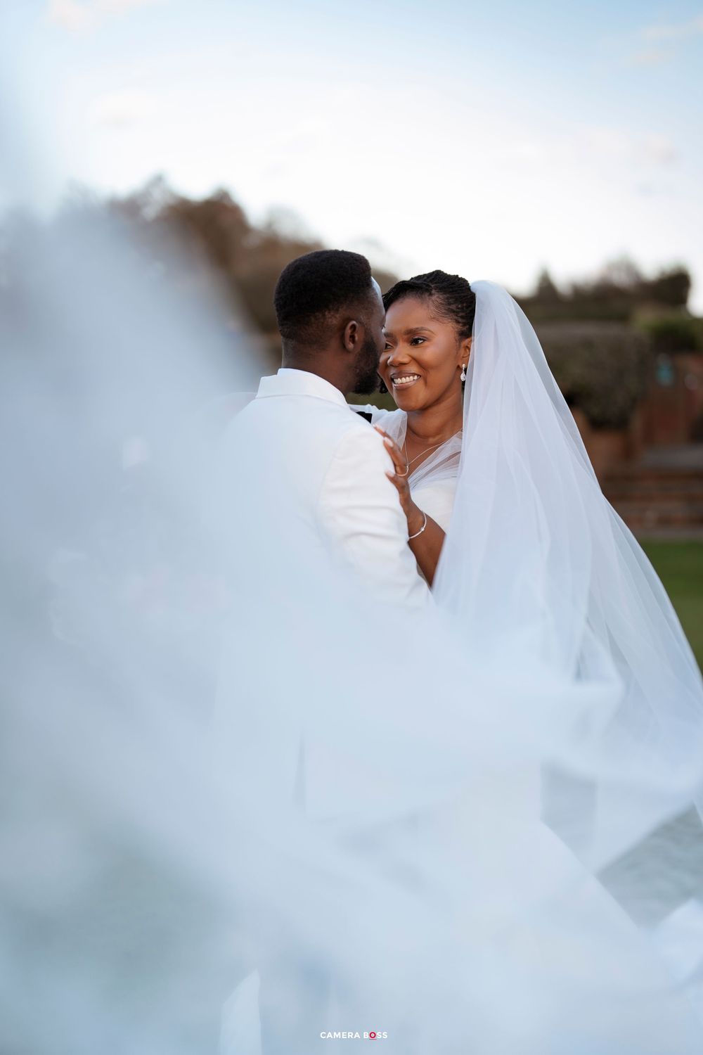 A joyful wedding moment captured through a veil as a couple shares an intimate embrace outdoors at sunset.