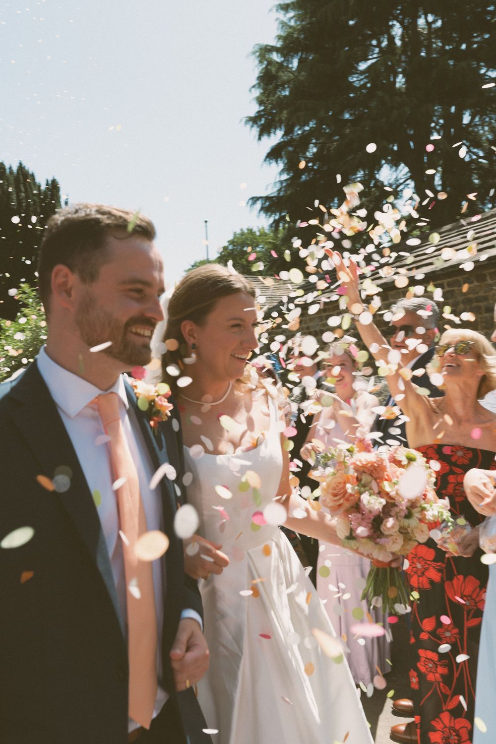 A happy couple celebrates their wedding day outdoors as guests shower them with white flower petals in the sunlight.