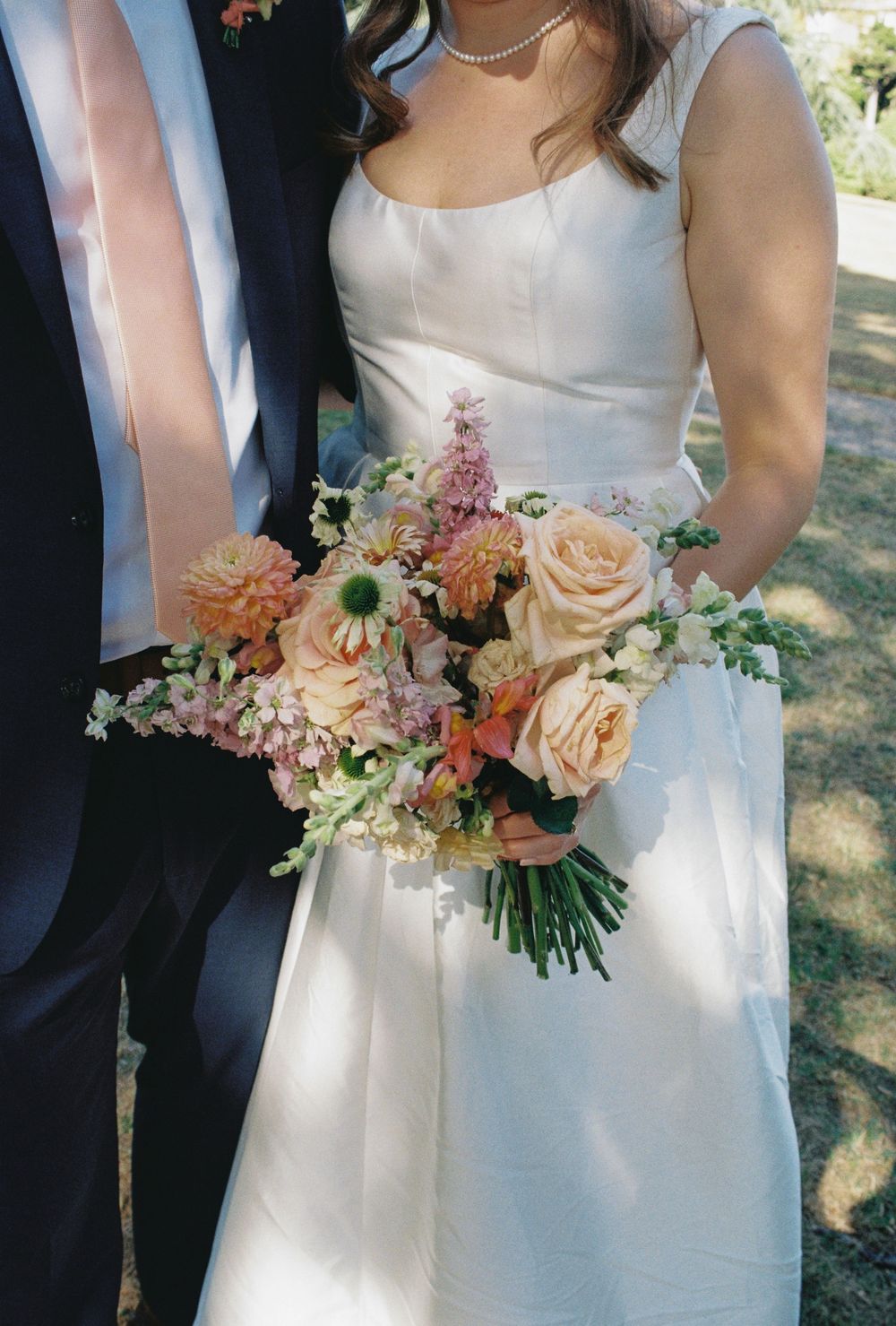 A wedding bouquet of peach roses and pink flowers held against a white bridal gown and dark suit.