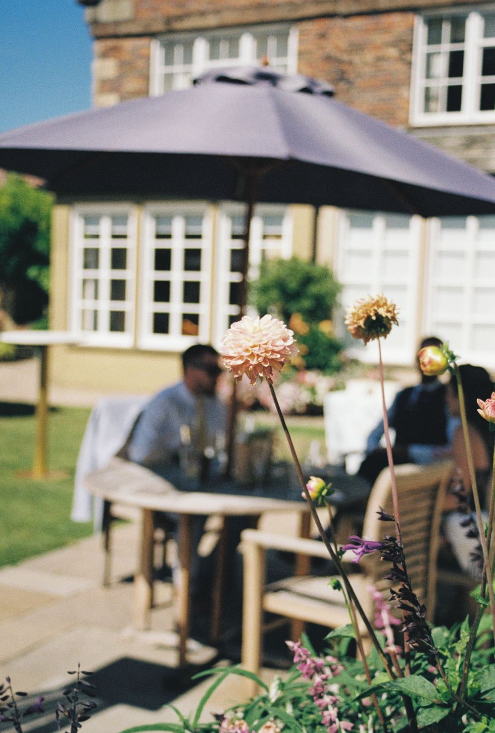 Outdoor patio seating area with umbrellas and flowering plants in foreground at a stone building on a sunny day.