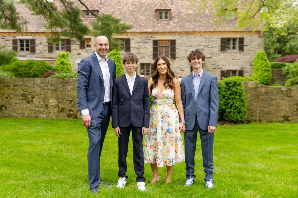 A group stands together on a manicured lawn in front of a stone country house on a sunny day.