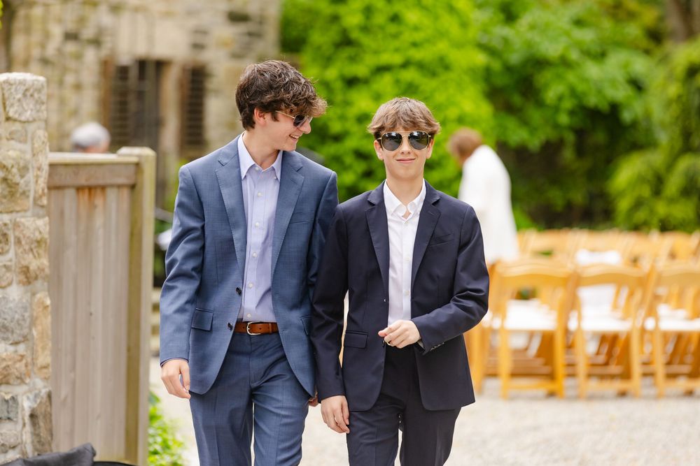 Three men in dark suits walk and chat along a stone pathway surrounded by greenery and outdoor event seating.