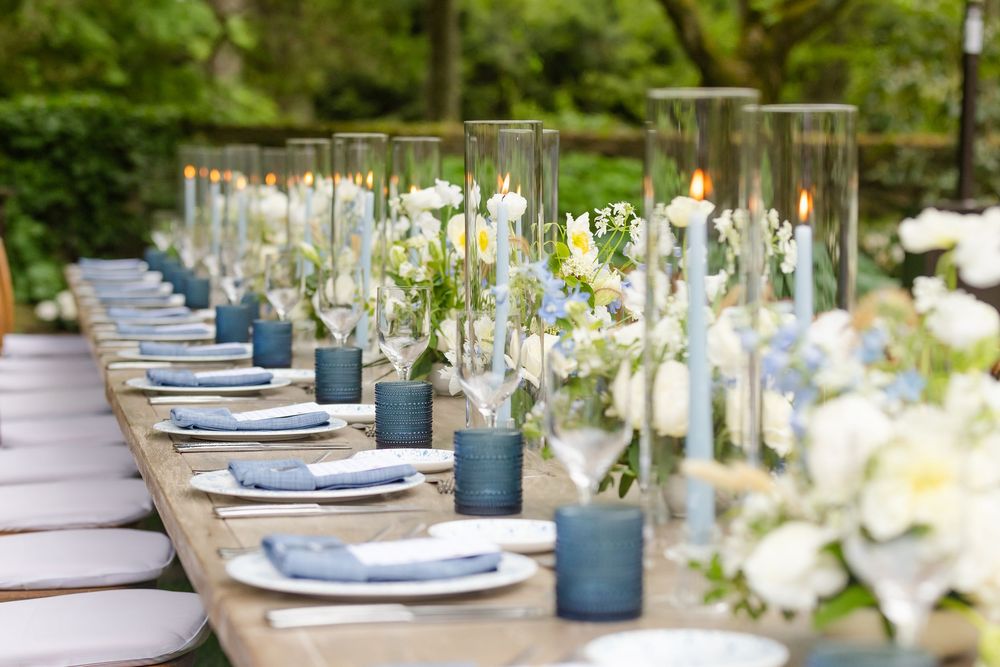 Elegant outdoor table settings with blue textured chargers, white flowers, and frosted glass vases along a rustic wooden table.