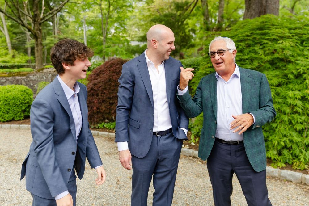 Three men in business suits laughing and having an animated conversation in a lush garden setting.