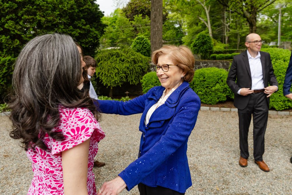 Guests in formal attire mingle and socialize at an outdoor gathering near lush green trees and shrubs.