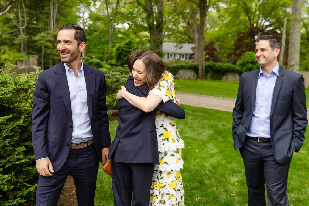 Two people in formal attire walk and laugh together through a lush green garden setting.