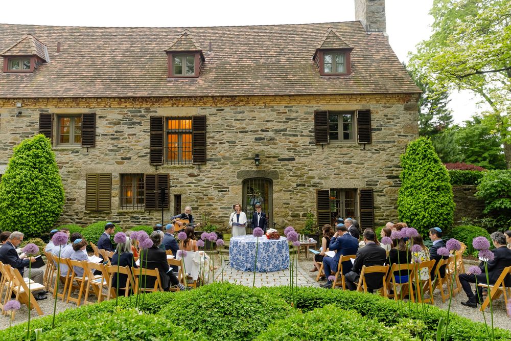 Multiple people gathered outside a rustic stone building with green landscaping during an outdoor event.
