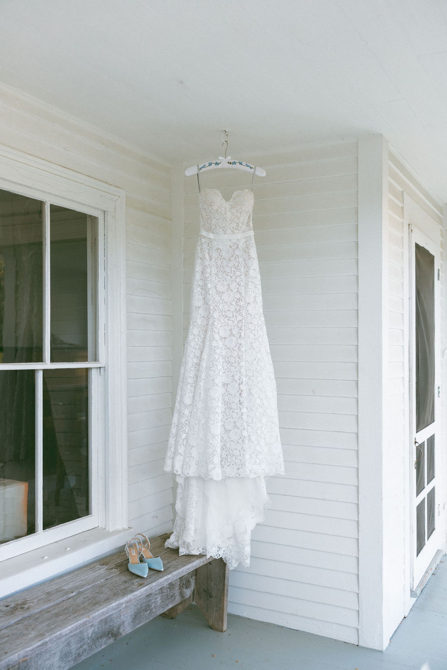 A white lace wedding dress hangs on display against a white wooden porch exterior.
