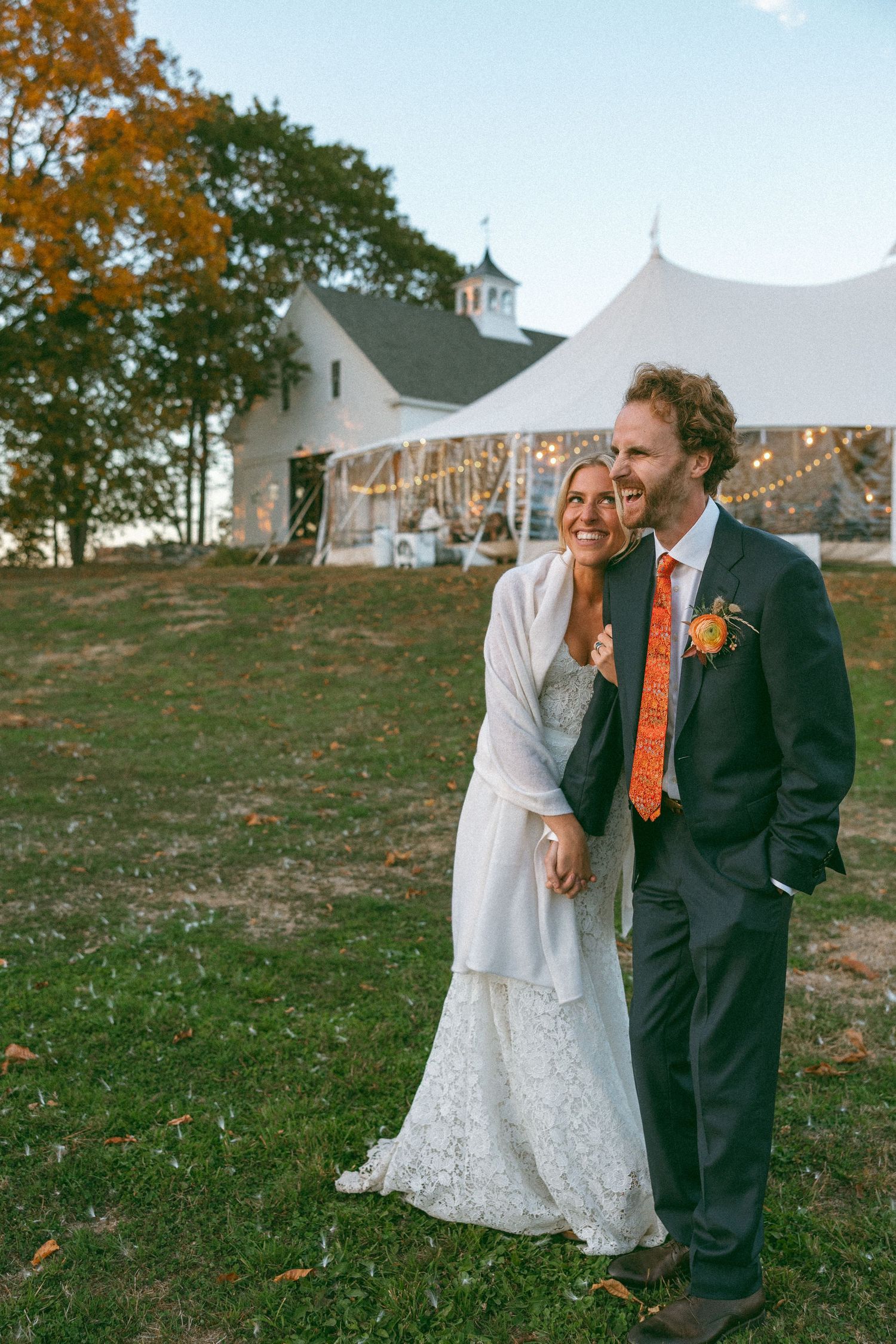 A couple shares a joyful moment outdoors with white tent and autumn foliage in the background.