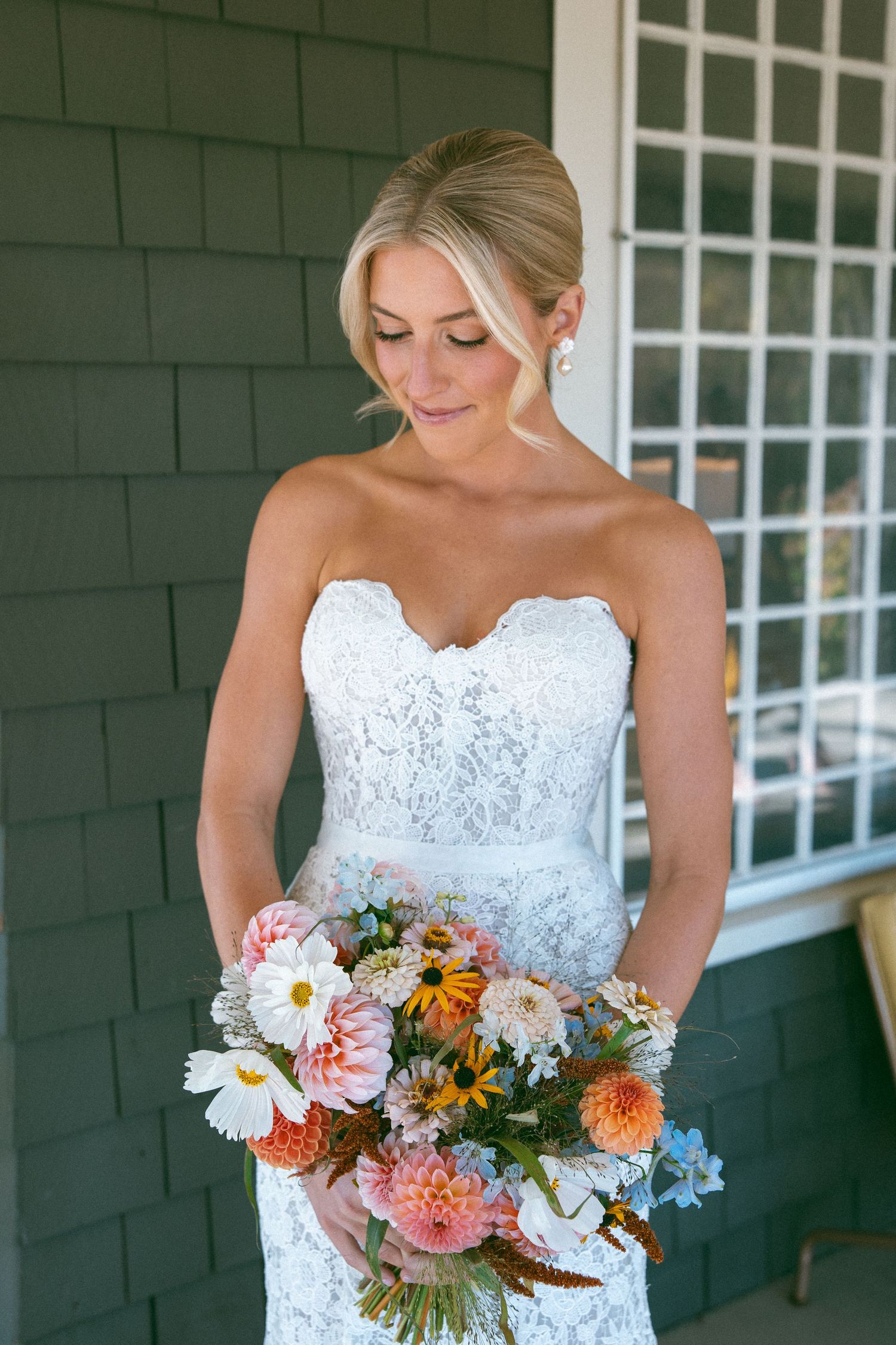 Bride in white lace dress holding a vibrant bouquet of seasonal flowers on a porch.