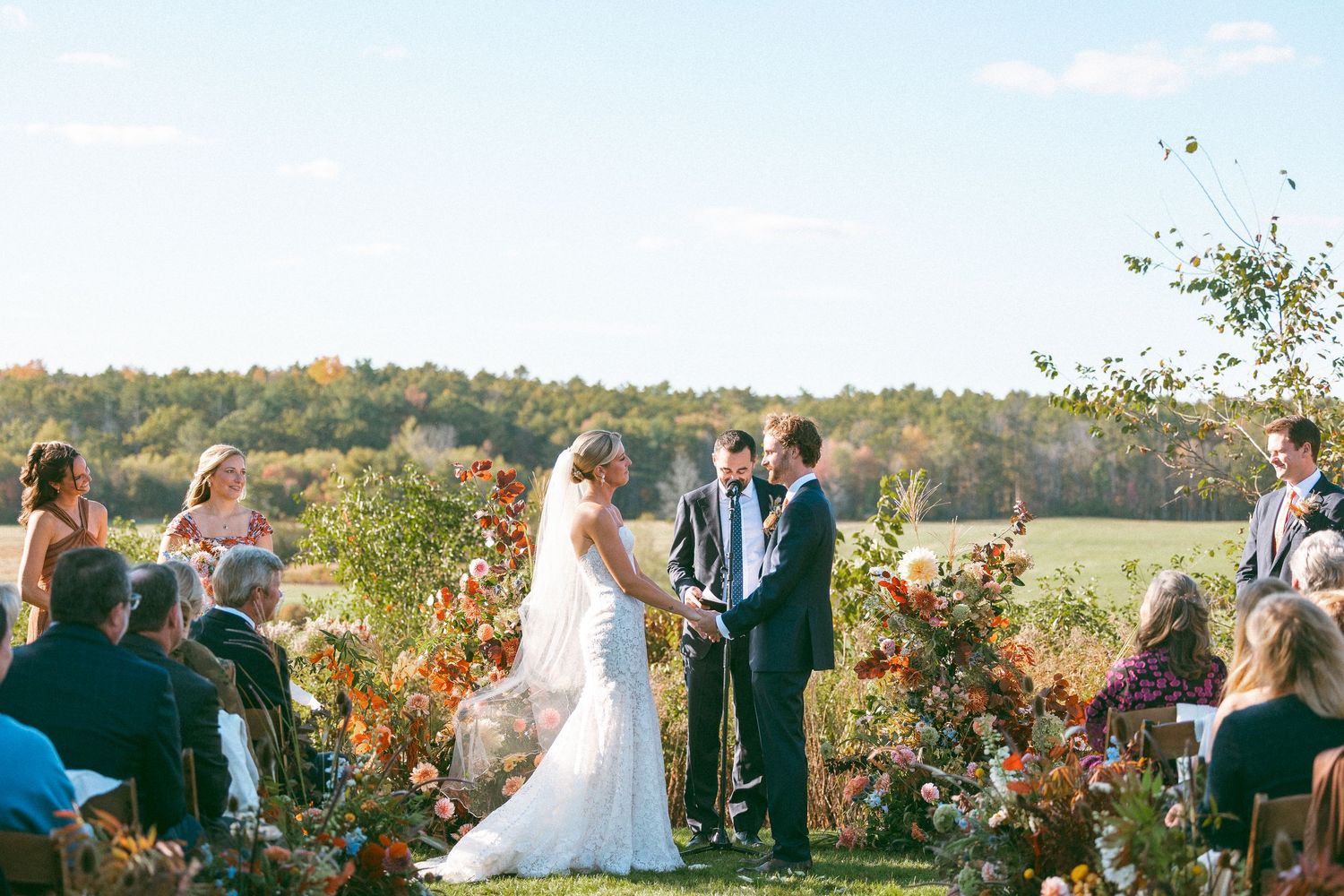 Wedding ceremony guests gathered outdoors on a hilltop during golden hour with fall foliage in the background.