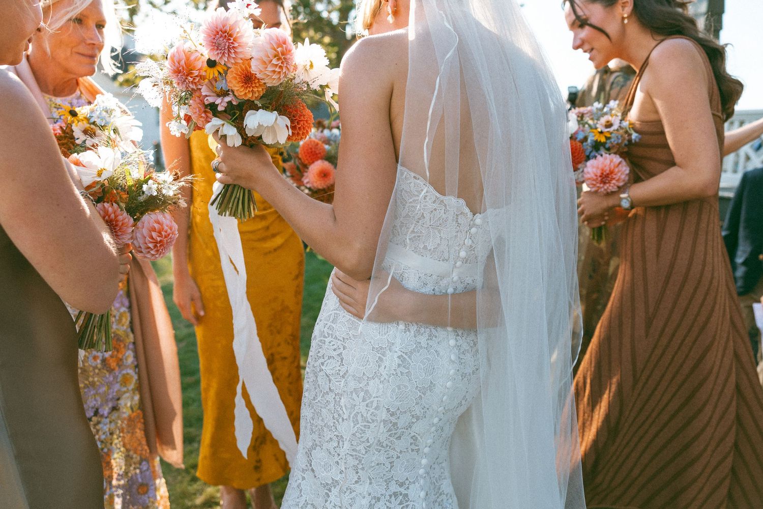 Wedding party members in autumn-colored dresses holding floral bouquets outdoors.