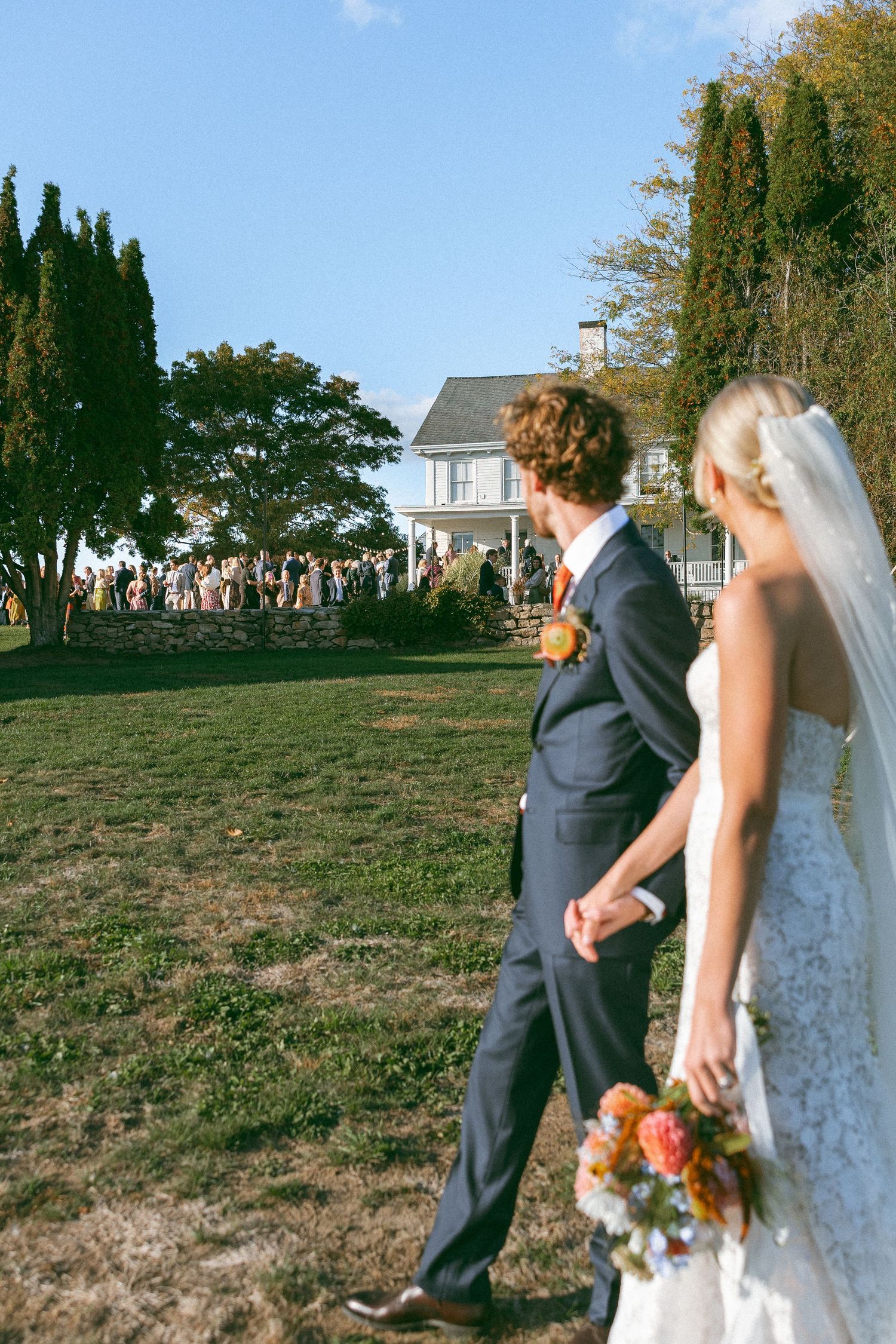 Wedding couple walking together on a grassy hill with white buildings in the background.