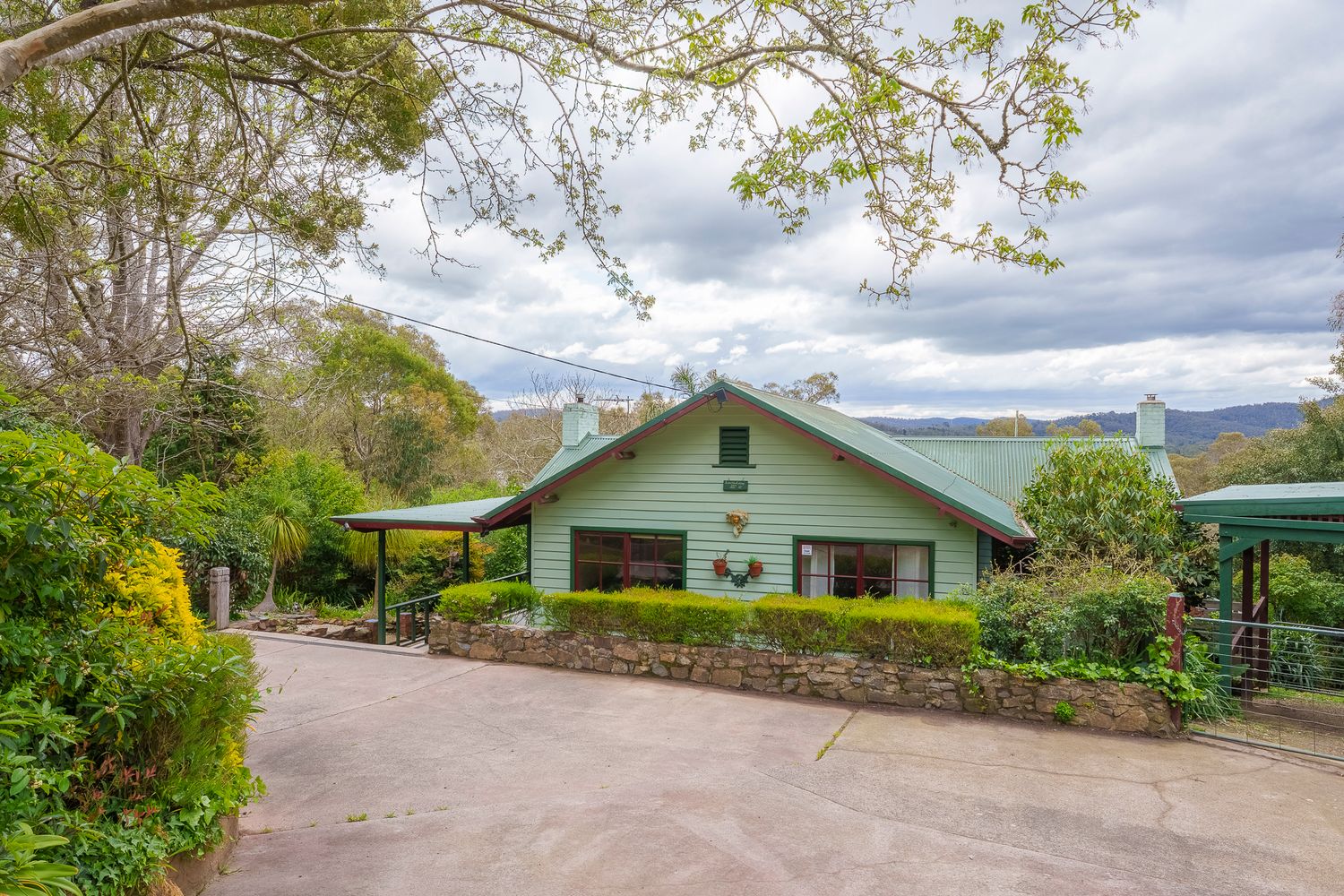 Charming green heritage cottage with a driveway, garden, and scenic mountain views under cloudy skies.