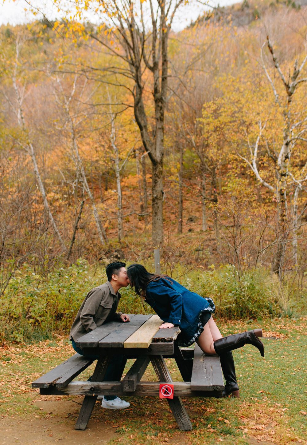 Two people laying on opposite sides of a wooden picnic table surrounded by autumn foliage in a park setting.