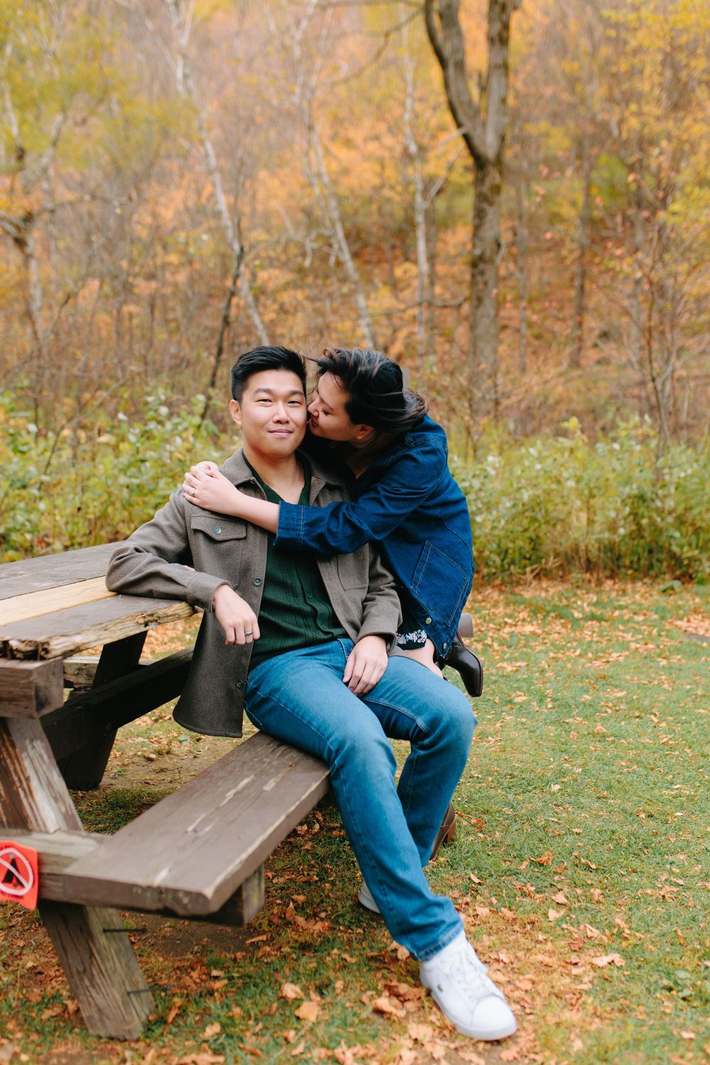 A couple shares an affectionate moment on a wooden picnic bench surrounded by autumn foliage in a park setting.