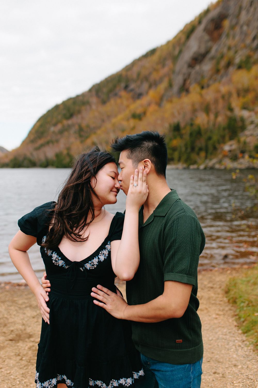 Couple shares intimate moments along a lakeside path with mountains in the background.