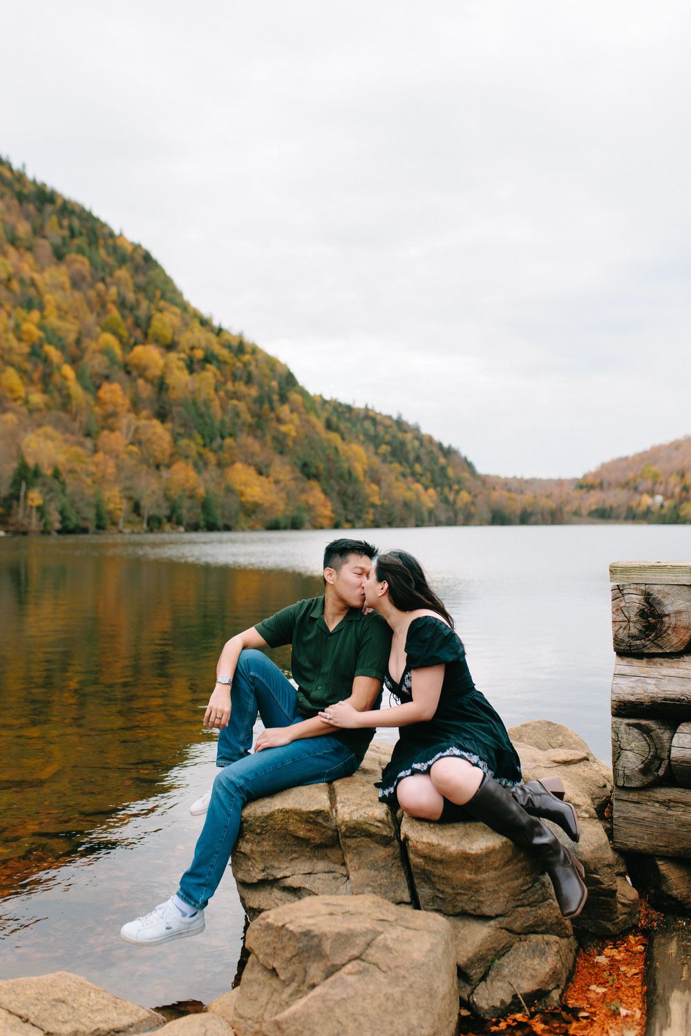Couple sharing a romantic moment on rocky shoreline with autumn foliage and lake views in the background.