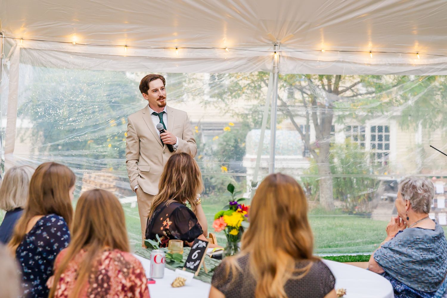 A speaker addresses guests under a white tent during an outdoor event with string lights and floral centerpieces.
