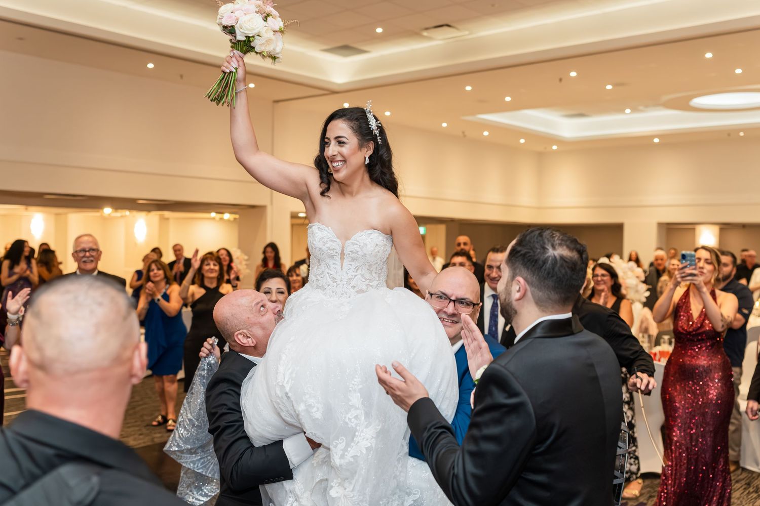 Wedding guests dancing and celebrating in an elegant reception hall with white decor and warm lighting.