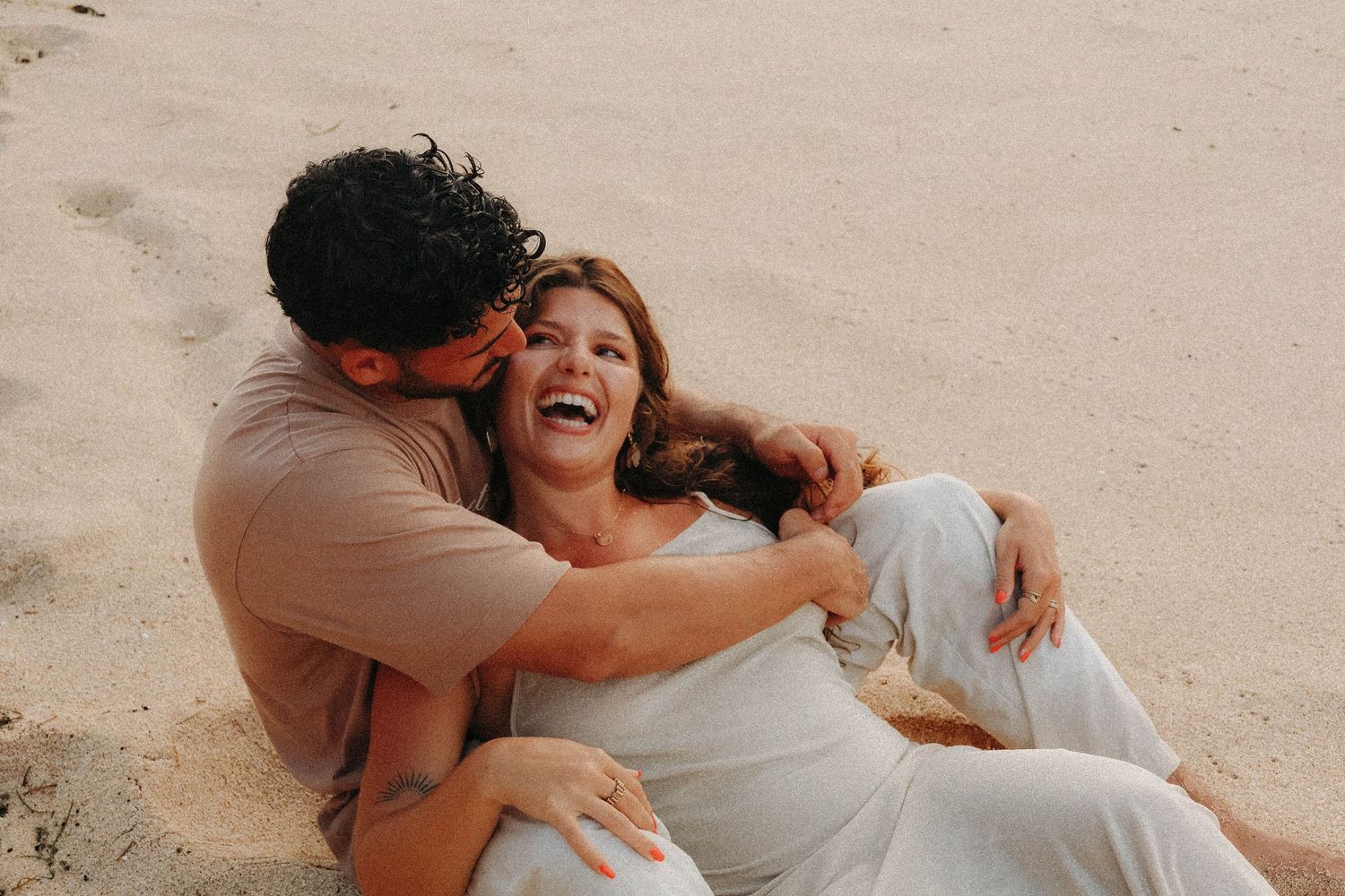 Laughing couple playfully cuddling on sandy beach, woman in white dress enjoying joyful moment together.