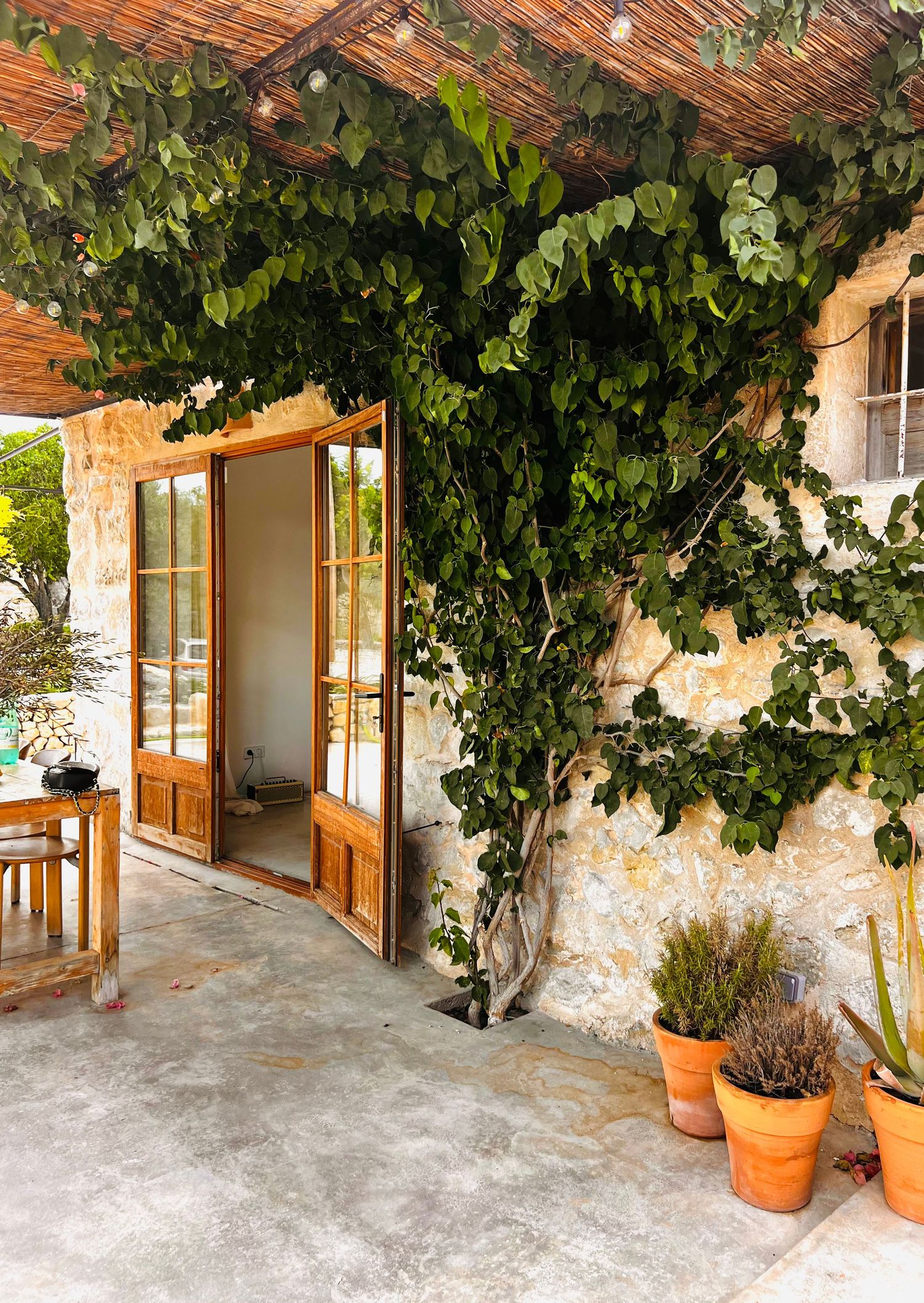 A rustic Mediterranean patio with stone walls, wooden pergola, and climbing vines creating a shaded outdoor living space.