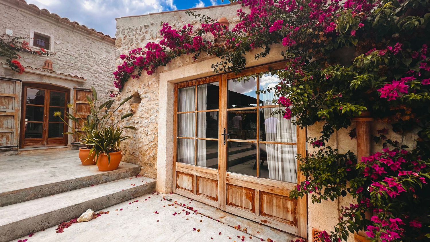 Vibrant pink bougainvillea flowers cascade over Mediterranean-style stone walls and wooden shutters in a charming courtyard.