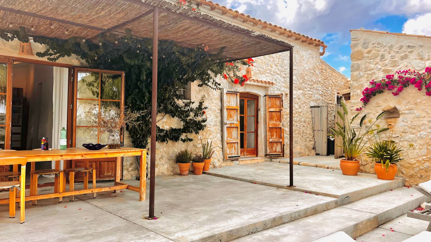 Panoramic view of a Mediterranean style courtyard with stone walls, terracotta pots, and wooden beams under blue sky.