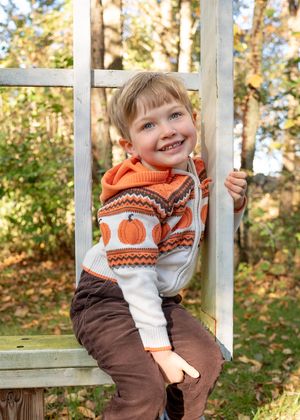 Child wearing an orange and brown patterned sweater sits playfully on a wooden porch railing in autumn.