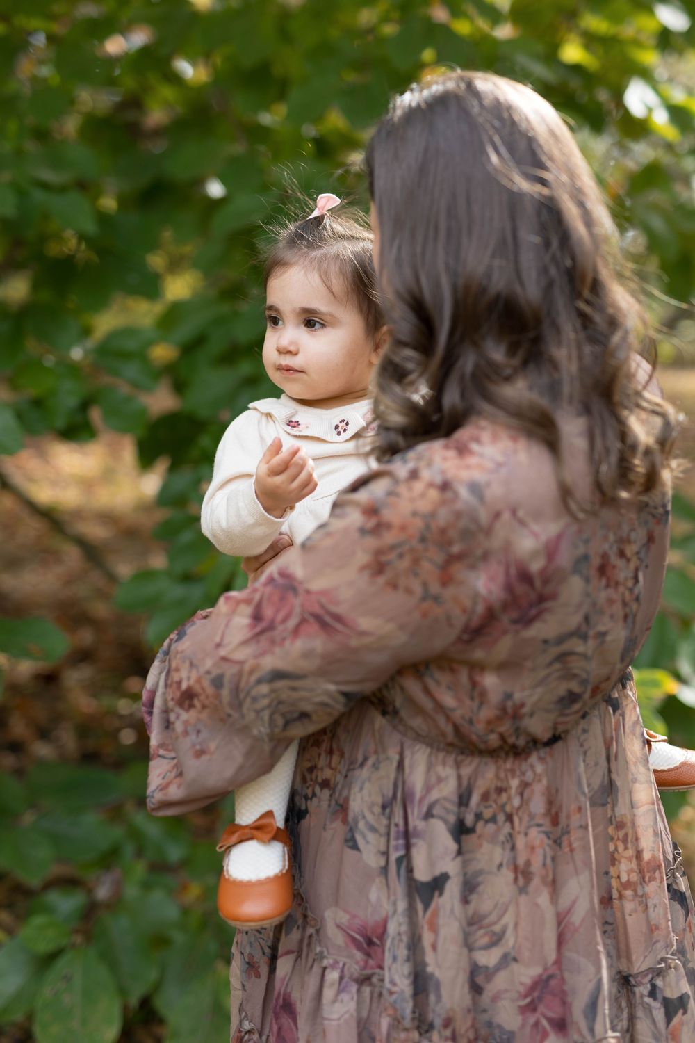 A parent in a floral dress holds a small child in white clothing against a green outdoor background.