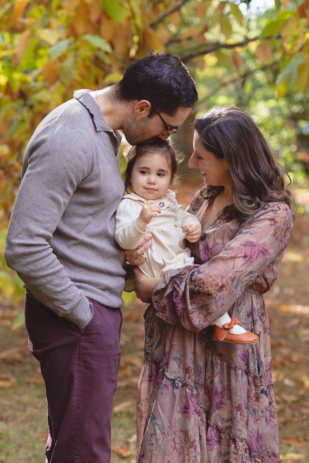 Family of three sharing tender moments together in an autumn forest setting with golden leaves in the background.