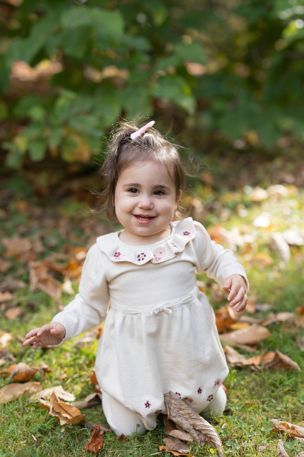 Small child in white dress sits on grass surrounded by autumn leaves in outdoor setting.
