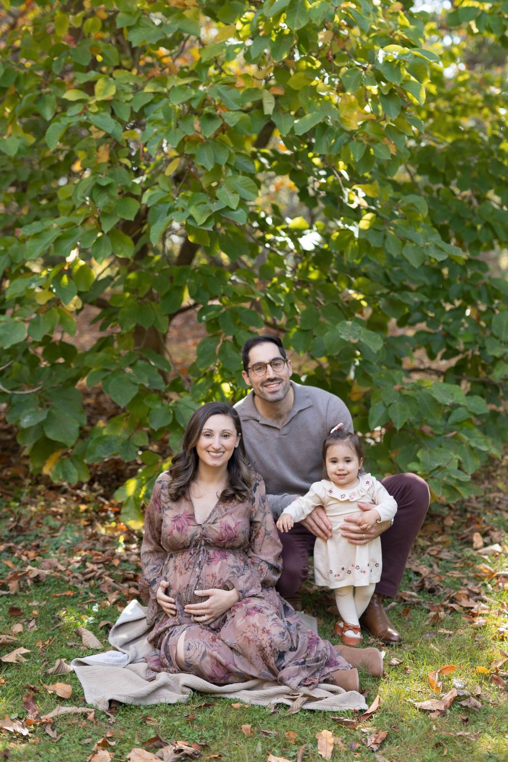 A family sits together on autumn ground beneath a leafy tree during a maternity photoshoot in nature.