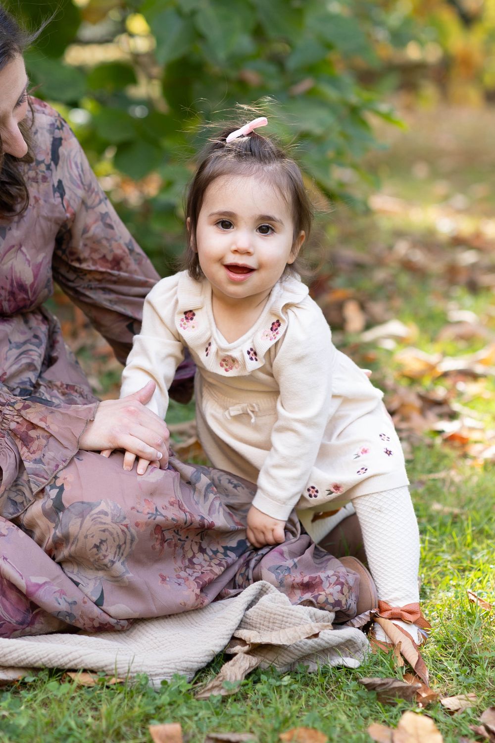A baby in a white sweater plays with autumn leaves on grass while sitting next to a brown blanket.