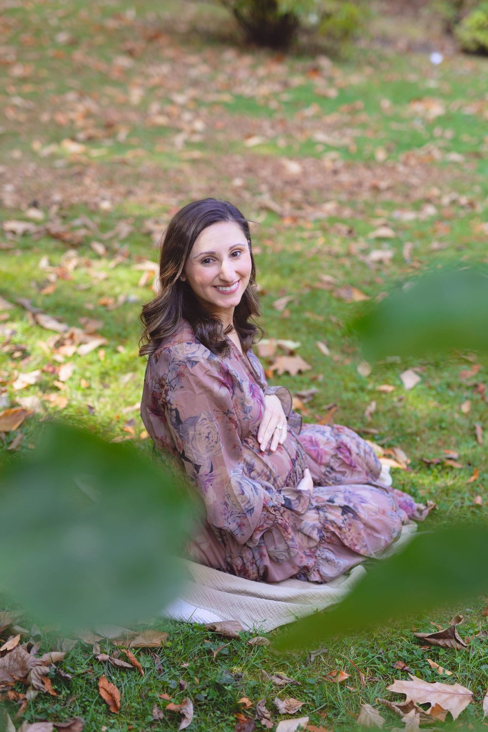 A woman in a floral dress reclines on a blanket surrounded by fallen leaves in an outdoor autumn setting.