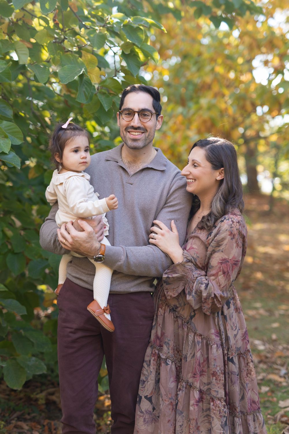 A family of three poses together outdoors against autumn foliage wearing coordinated brown and mauve outfits.