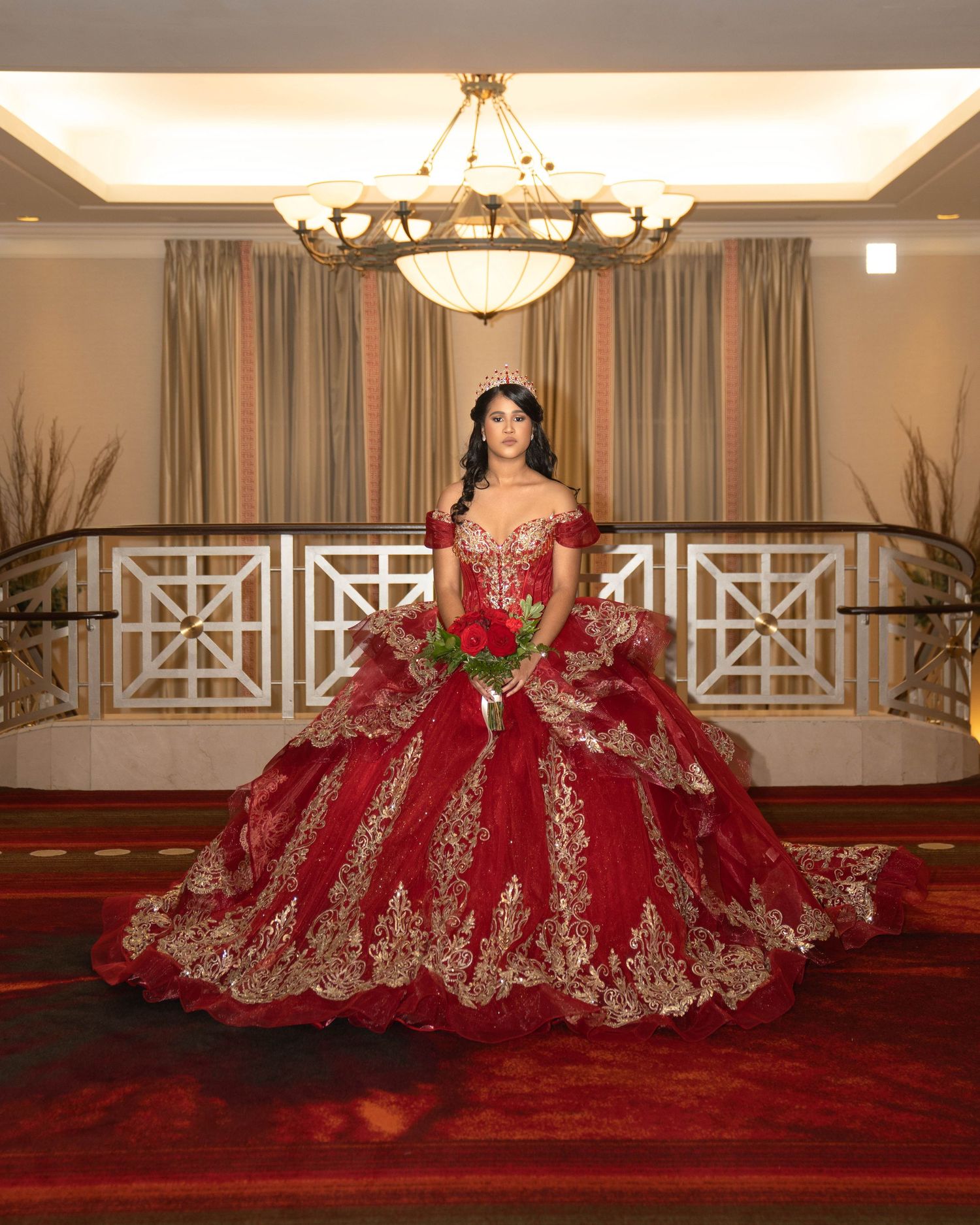 A stunning red and gold quinceañera dress displayed in multiple angles on a red carpet in an elegant ballroom setting.