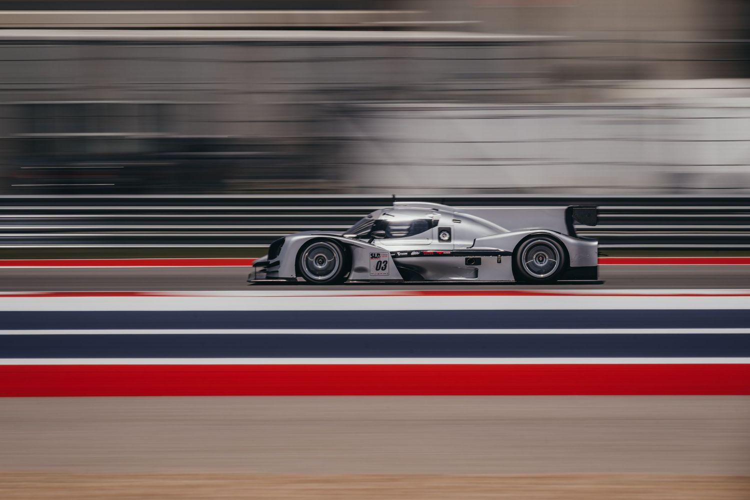 Racing car speeds along a racetrack with motion blur against cloudy sky and mountains in the background.