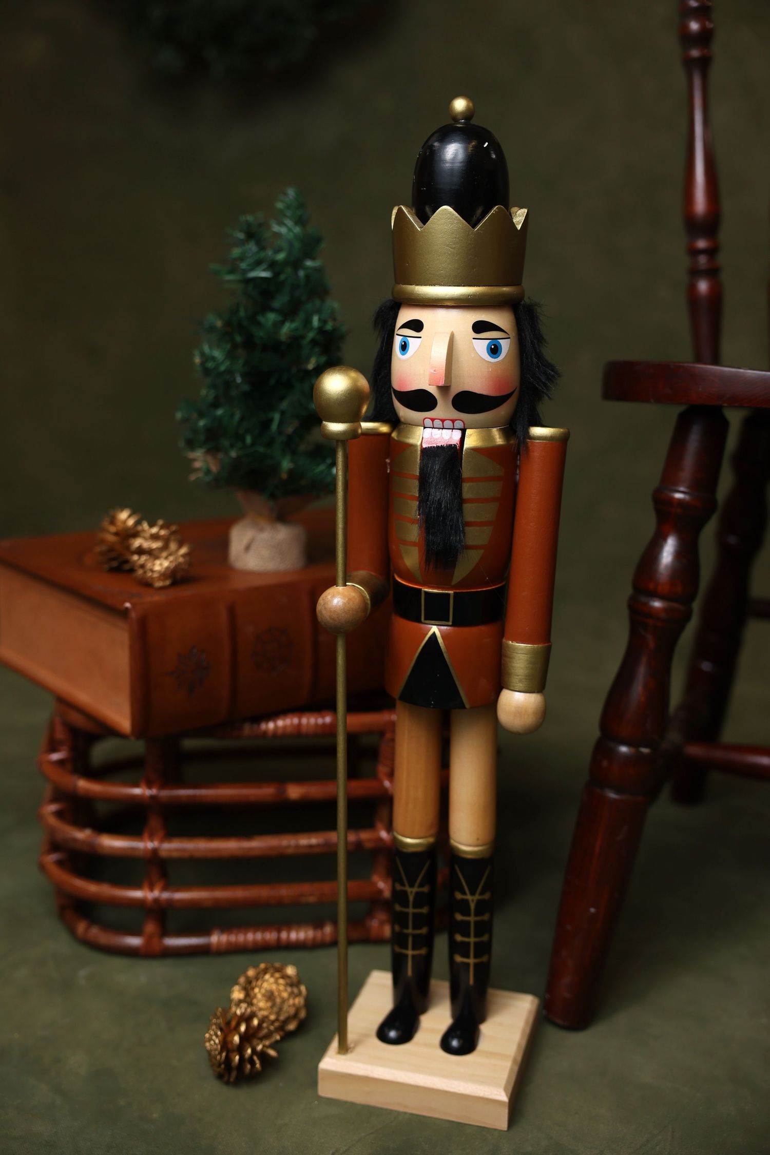 Traditional wooden nutcracker soldier figurine with gold crown stands next to stacked books and pine cones in festive holiday scene.