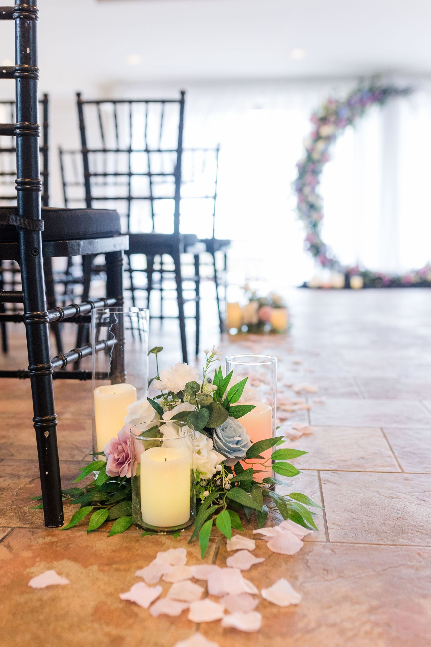 Pink rose petals scattered on a stone floor with black chairs and glowing candles create an elegant wedding reception setting.