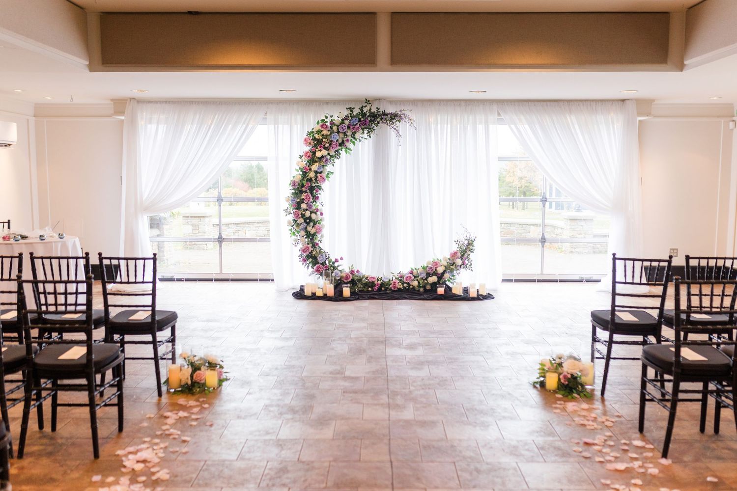 Elegant white wedding venue interior with dark wooden chairs and natural lighting through large windows.