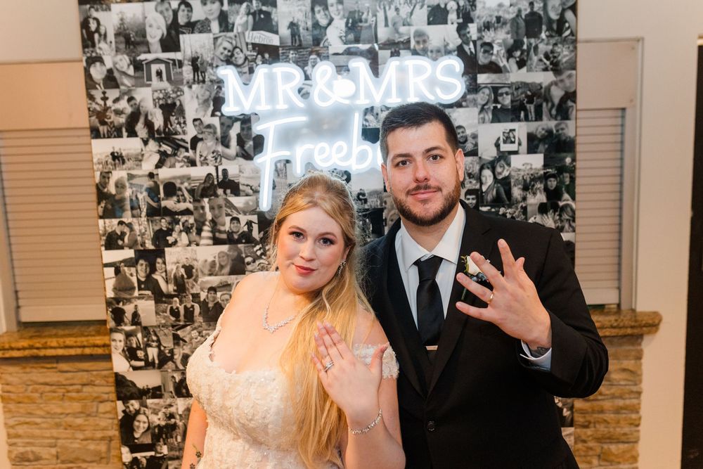 A couple poses together at their wedding reception in front of a black and white photo wall collage.