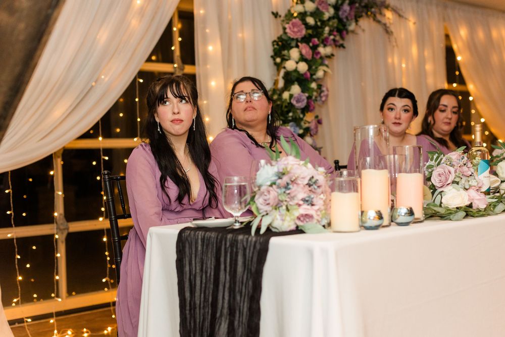 Wedding reception head table decorated with white linens, floral arrangements and twinkling lights in an elegant indoor venue.