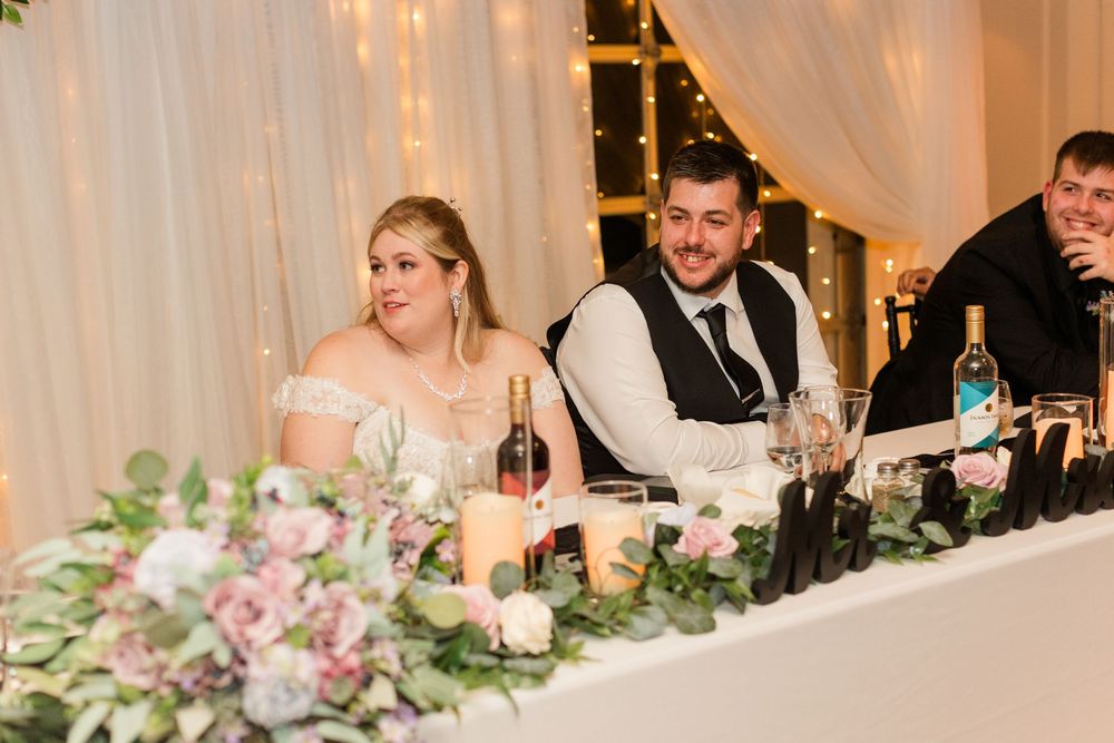 Wedding party members seated at a long head table decorated with floral centerpieces and soft lighting.