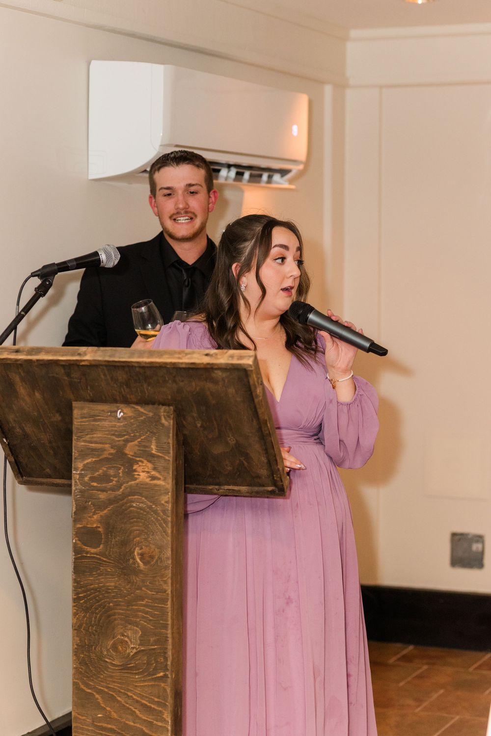 A speaker in a lavender dress delivers remarks at a wooden podium during an indoor event.