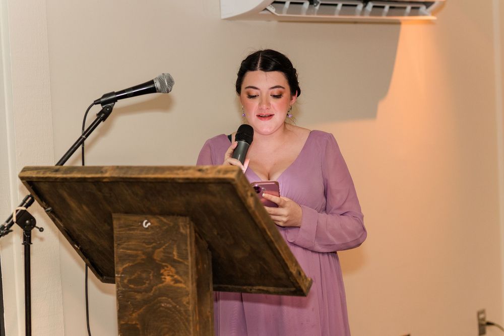 A person in a purple dress speaks at a wooden podium with a microphone during an evening celebration.