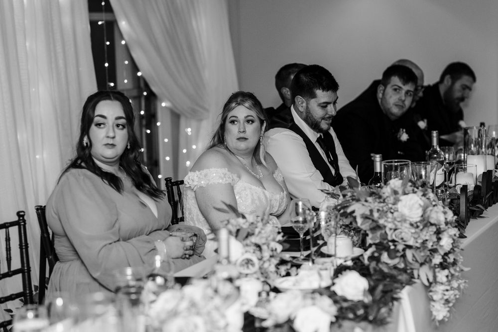 Wedding party members seated at an elegant reception table with floral centerpieces and twinkle lights in black and white.