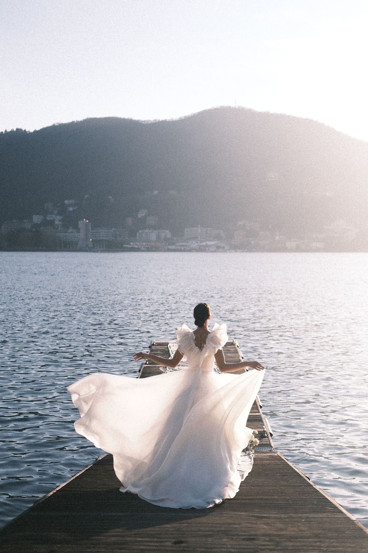 Bride in a white flowing gown stands on a boat on a large lake with mountains